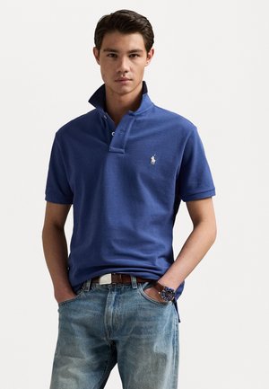 Blauwe katoenen polo met een verhoogde kraag, een knoopsluiting met twee knopen en een geborduurd logo. G gedragen met lichtblauwe denim en een bruine riem.