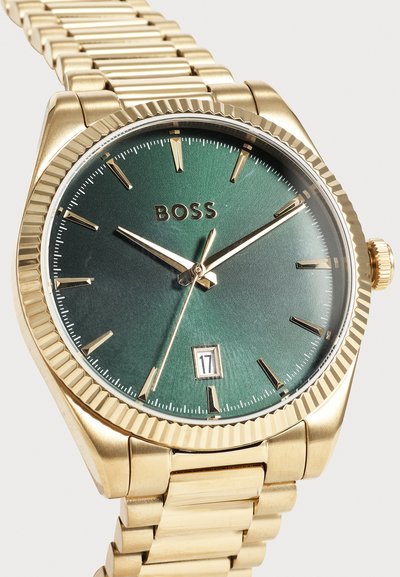 Montre-bracelet en or avec cadran vert, index horaires et aiguilles dorés, affichage de la date à 6 heures et bracelet en maillons métalliques indiquant l’heure 10:09:36.