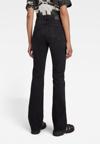 Osoba v černých bootcut džínách a černých sandálech, stojící s zády otočenými k fotoaparátu na jednoduchém bílém pozadí.