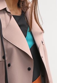 Manteau double boutonnage rose clair avec un col cranté, boutons noirs et un haut à motif contrasté teal et noir en dessous. Texture lisse.