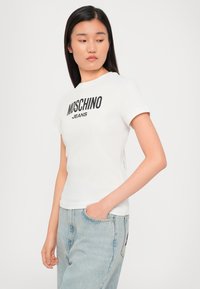 White fitted T-shirt with short sleeves, featuring a black "MOSCHINO" logo and "JEANS" text, paired with light blue denim jeans.