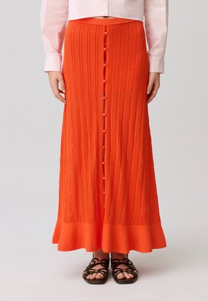 Gonna lunga arancione in maglia con bottoni sulla parte anteriore, indossata con sandali neri con cinturini e una camicetta rosa chiaro a maniche lunghe.