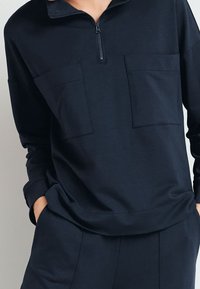 Marineblauwe pullover met een kraag met kwartrits, twee borstzakken en een losse pasvorm. Zacht materiaal met een gladde textuur en lange mouwen.