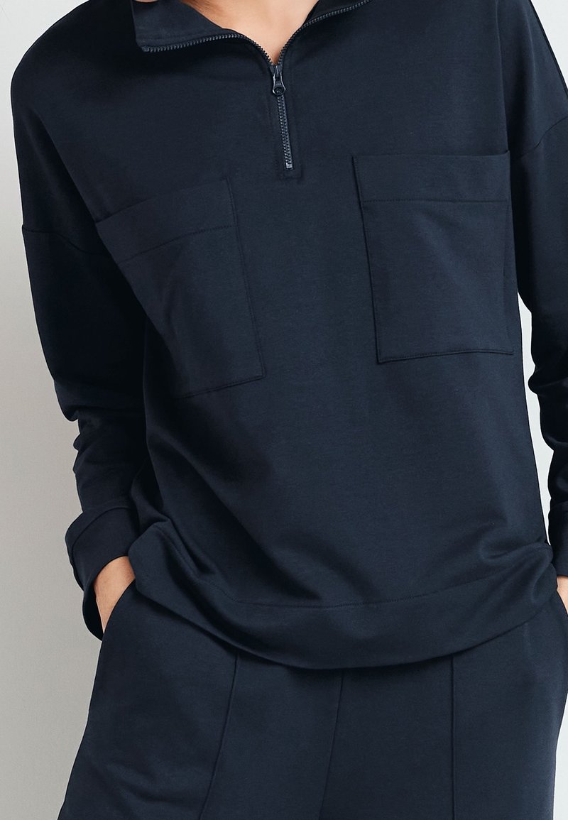 Marineblauwe pullover met een kraag met kwartrits, twee borstzakken en een losse pasvorm. Zacht materiaal met een gladde textuur en lange mouwen.