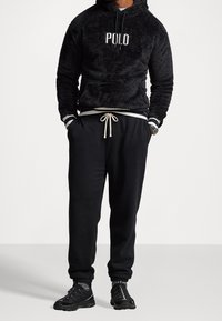 Man som bär en svart fluffig Polo-hoodie, svarta sweatpants med vit snörning, svarta sneakers och grå strumpor, stående med händerna i fickorna.