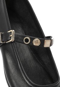Zapato de cuero negro con un diseño plano. Presenta una correa decorativa con acentos metálicos, incluyendo un remache circular y un detalle cuadrado texturizado.
