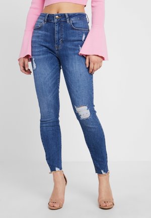 Frau trägt hoch taillierte blaue, zerrissene Skinny-Jeans, ein pinkes langärmliges Cropped-Top und beige Knöchelriemen-Absätze, steht vor einem schlichten Hintergrund.