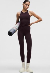 Bordowy sportowy top na cienkich ramiączkach, dopasowane legginsy i białe obuwie. Pod jedną ręką trzymana jest wielobarwna mata do jogi.