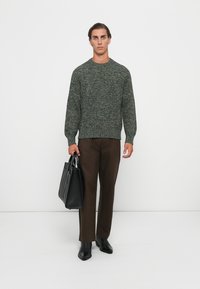 Green textured knit sweater with a round neck, paired with brown trousers and black ankle boots. A black bag is held in the right hand.