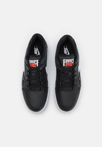 Sapatilhas de basquetebol sintéticas negras com parte superior perfurada, swoosh branco da Nike e logótipo vermelho/preto na língua. Sola de borracha branca.