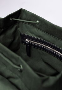 Interno di una borsa tote in tessuto verde con una tasca con zip in pelle nera. Presenta una chiusura con cordino e una texture liscia e opaca.