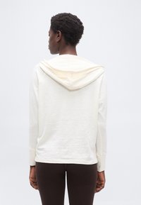 Άτομο με κοντά κατσαρά μαλλιά που φοράει φούτερ με κουκούλα σε εκρού χρώμα και σκούρο καφέ παντελόνι, από πίσω σε απλό φόντο.