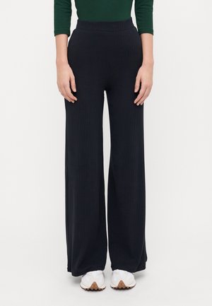 Persoon draagt zwarte wijde ribbroek, groene top met lange mouwen en witte sneakers, staand tegen een effen witte achtergrond.