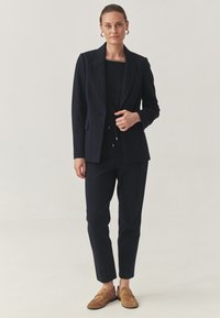 Sacou bleumarin cu rever structurat, asortat cu un top negru și pantaloni bleumarin croiți. Loafers maro completează ținuta. Textură netedă pe toată suprafața.