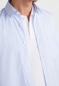 Ljusblå randig knapp-up skjorta med klassisk krage, tillsammans med en vit polo under. Har en bröstficka och en slät textur.