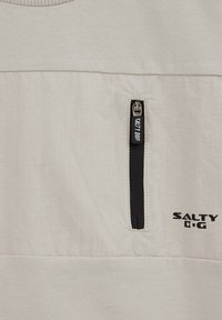 Šviesiai smėlio spalvos audinio panelė su vertikaliu juodu užtrauktuku, pažymėtu "SALTY DOG", ir šalia esantys juodu atspausdinti žodžiai "SALTY DOG".