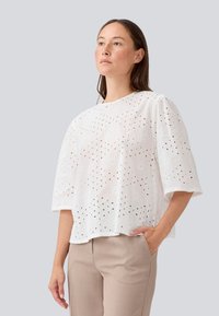 Blouse en coton blanc avec des détails floraux brodés, présentant des motifs découpés, des manches courtes et une coupe carrée. Portée avec un pantalon beige.