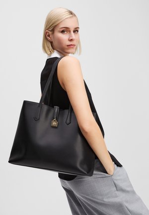 Borsa tote in pelle nera con due manici, design minimalista, caratterizzata da un accento a forma di lucchetto dorato quadrato. Texture liscia e forma strutturata.