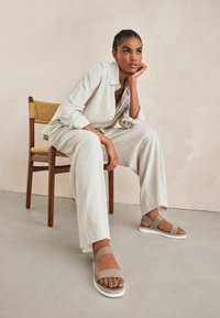 Bruna och beige sandaler med plattformssula och justerbara ankelsnodder. Modellen bär en ljusbeige linneoutfit och sitter på en trästol.