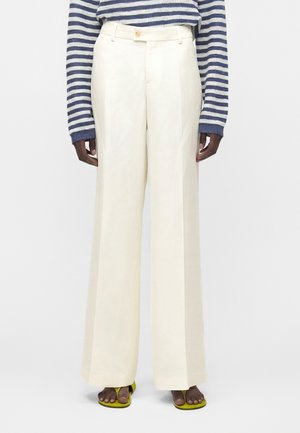 Άτομο που φοράει κρεμ παντελόνι με ψηλή μέση και φαρδιά μπατζάκια, πουλόβερ με ναυτικό και λευκό ριγέ μοτίβο και σανδάλια λαχανί, στέκεται μπροστά σε απλό φόντο.