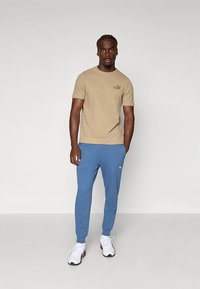 Beige t-shirt med korte ærmer og logo, kombineret med blå joggingbukser. Hvide sportssko med sorte og røde detaljer bæres.