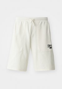 Shorts de couleur crème fabriqués à partir d'un tissu doux, dotés de deux grandes poches latérales et d'un logo noir "Converse All Star" sur la jambe droite.
