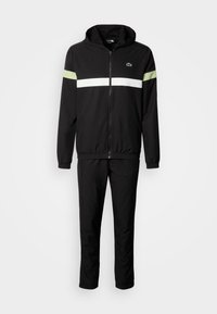 Musta verkkaritakki, jossa on vetoketjullinen takki ja valkoiset sekä vaaleanvihreät raidat olkapäillä. Pehmeää kangasta, sisältää hupun ja joustavat hihansuut.