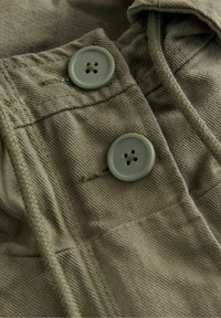 Gros plan de deux boutons verts cousus sur un tissu vert olive avec des coutures visibles et des plis texturés.