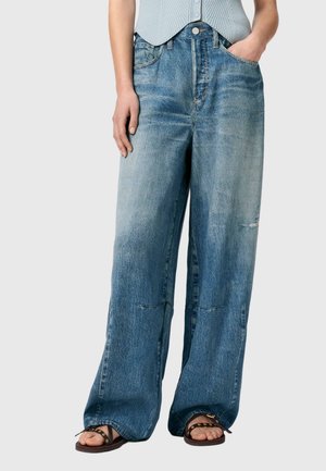 Personne portant un jean bleu délavé et ample avec une main dans la poche, associé à des sandales marron à bouts ouverts et un haut sans manches bleu clair.