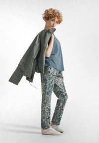 Casuele outfit met een losse blauwe top, groene jas en een broek met een blauw-beige botanisch patroon. Witte sneakers maken de look compleet.