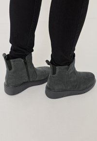 Harmaat mokkasaappaat nilkkoihin, joissa on sivuvetoketjut, teksturoitu yläosa ja mustat kumipohjat. Suunnittelussa on minimaalisesti ompelua ja joustavia inserts.