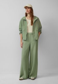Svijetlozelena zip-up jakna, bež majica, široke svijetlozelene hlače, bež kapa i bež sandale. Glatka tekstura, opušteni kroj i slojeviti dizajn.