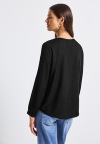 Blusa nera a maniche lunghe con scollo a barca e polsini con bottoni, abbinata a jeans in denim azzurro chiaro, vista da dietro.