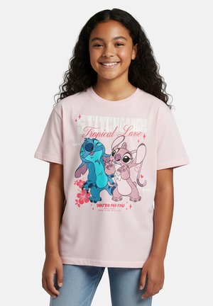Smiling girl with long curly hair wearing a light pink Stitch and Angel “Tropical Love” graphic T-shirt and blue jeans.