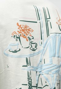 Lichtgekleurde t-shirt met een lineair ontwerp van een tafel met een vaas met bloemen en koffiekopjes in blauw, groen en oranje.