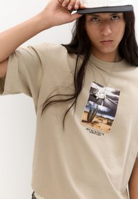 Beige kortærmet t-shirt med et grafisk print, der viser en ørken-scene, kaktusser og tekst. Tekstureret stof, afslappet pasform, synlige sømme.