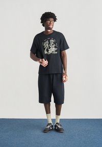 Μαύρο γραφικό t-shirt με κίτρινο σχέδιο, συνδυασμένο με μαύρο σορτς. Το μοντέλο φοράει ανοιχτόχρωμες κάλτσες και μαύρα παπούτσια Nike.