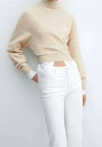 Maglione beige cropped con colletto alto e bordo a coste, abbinato a jeans bianchi aderenti con bottoni laterali e dettaglio con aperture.