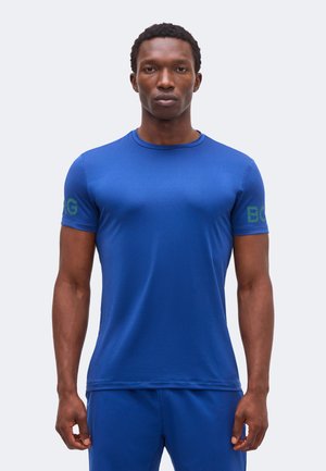 Mann iført en tettsittende blå kortermet skjorte og matchende blå bukser, stående mot kameraet mot en ensfarget bakgrunn.