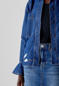 Giacca di jeans con vestibilità leggermente allentata, colore blu scuro, taschino sul petto e strappi sulla parte inferiore anteriore. Abbinata a un top blu navy.