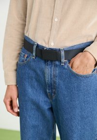 Svart nylonbälte med en rektangulär spänne, bärs över en ljusbeige skjorta och blå denimjeans, som visar fickor och en blekt textur.