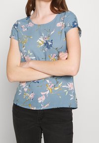 Kvinde med korslagte arme iført en blå blomster- kortærmet top og sorte bukser, stående mod en ensfarvet lys baggrund.