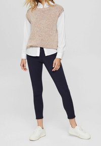 Stickad beige tröja med flerfärgade fläckar, lagd över en vit skjorta med knappar. Matchad med mörka skinny jeans och vita sneakers.