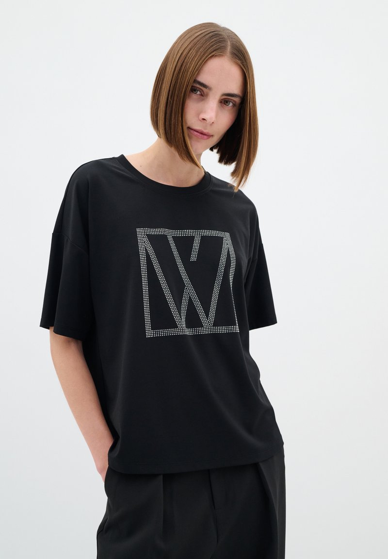 T-shirt noir oversize à manches courtes, avec un imprimé logo géométrique en strass argenté sur le devant. Texture de tissu douce et lisse.