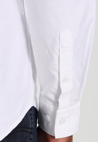Camicia bianca con tessuto testurizzato, con polsini dritti e due bottoni trasparenti, e un design di colletto classico.