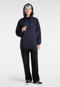Sweat à capuche navy avec une poche frontale et un cordon de serrage, arborant un design graphique teal. Assorti avec un pantalon noir et des baskets noir et blanc.