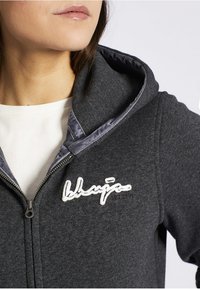 Sudadera con capucha gris con cremallera negra a contraste, capucha forrada y logo blanco bordado en el pecho. Tejido suave y texturizado.