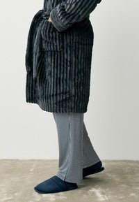 Gray ribbed robe with a tie closure and pockets, paired with striped pants in blue and white, and navy slippers on a beige floor.
