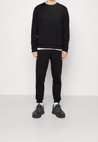 Man som bär en svart sweatshirt med subtil "BOSS"-logotyp, svarta joggerbyxor, vita strumpor och svarta sneakers med reflektiv snörning, står upp.