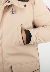 Veste isolée beige avec une fermeture éclair à l'avant, des poignets élastiques et une poche latérale. Présente un col texturé et des surpiqûres noires.